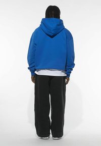 Blå oversized hoodie med hætte, der har dropped shoulders og en afslappet pasform, båret over en hvid skjorte og parret med sorte vide bukser.