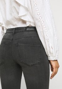 Graue Jeans aus Denim mit figurbetontem Schnitt, ausgestattet mit zwei Gesäßtaschen und einem schwarzen Lederetikett am Bund. Weißes Hemd mit Lochstickerei.