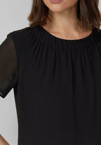 Vrouw draagt een zwarte top met een gerimpelde halslijn en doorschijnende korte mouwen, afgebeeld van nek tot midden torso tegen een effen achtergrond.