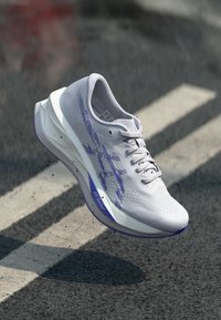 Λευκό αθλητικό παπούτσι με πλέγμα στο πάνω μέρος, ανοιχτό μωβ λεπτομέρειες, υφή επιφάνειας και ευαίσθητη σόλα. Ελαφρύς σχεδιασμός με σύστημα δέσης κορδονιών.