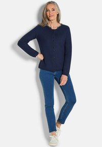 Frau mit grauem Haar, die eine marineblaue Knopfpullover-Jacke, blaue Jeans und weiße Sneaker trägt, steht mit einer Hand auf der Hüfte vor einer weißen Wand.