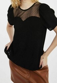 Svart topp med en nät-yoke med prickigt mönster, korta puffärmar och avslappnad passform. Matchas med en brun faux läderkjol.
