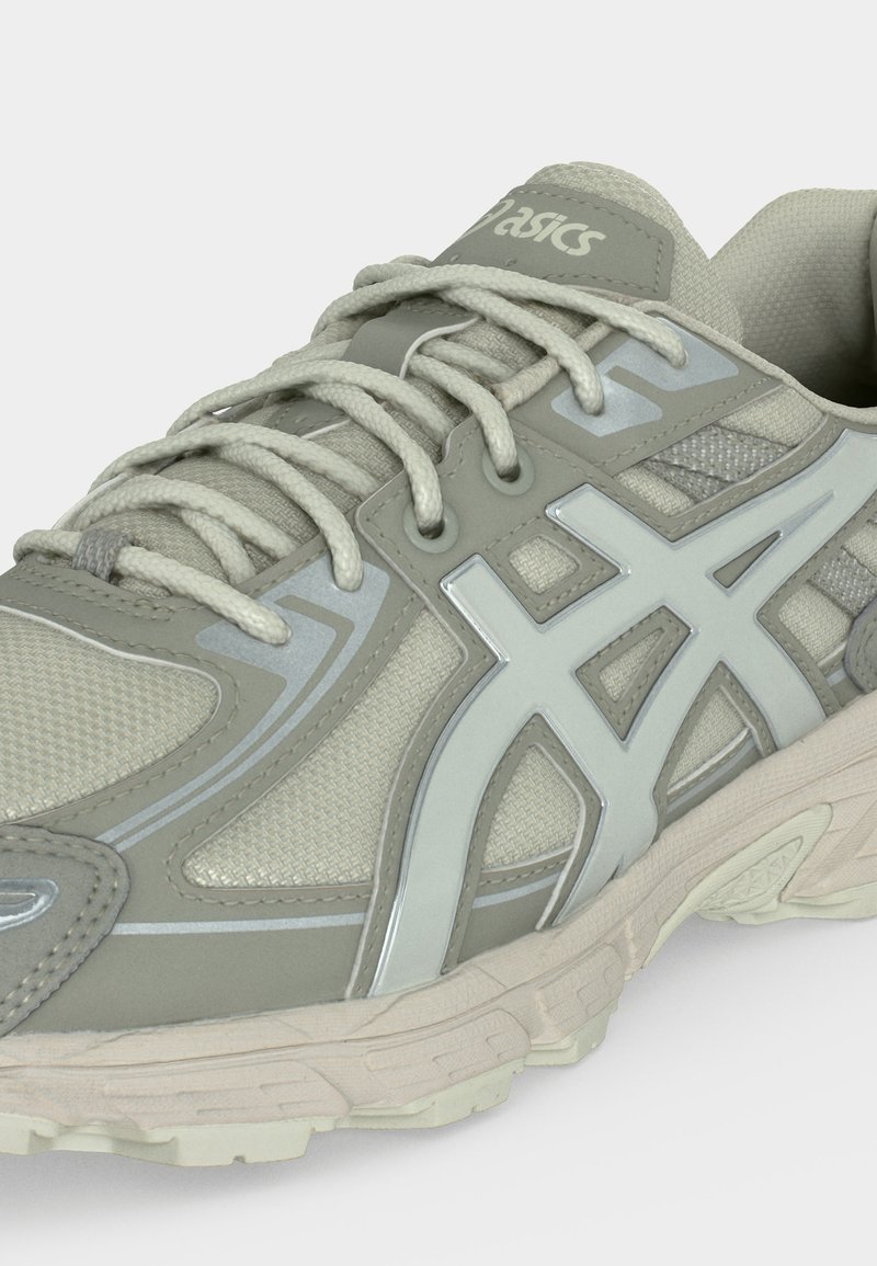 Zapatilla de running Asics de color gris claro con tejido de malla texturizada, cuello acolchado, cordones y suela de goma gruesa con dibujo detallado.