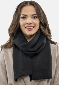 Sort strikket tørklæde med en blød tekstur, der har et bredt, draperet design og ribbede ender, båret over en lysefarvet frakke.