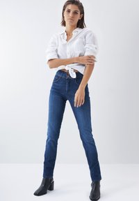 Jeans de tiro alto en azul oscuro, con corte ajustado, hechos de mezclilla, que presentan tres botones frontales y costuras sutiles. Combinados con una camisa de lino blanca.