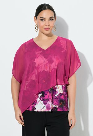 Frau trägt ein pinkes, durchsichtiges florales Top über einer bedruckten Bluse mit lila Blumen und schwarzen Hosen, stehend vor weißem Hintergrund.