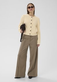 Krēmkrāsas ribota jaka ar melnām pogām, saskaņota ar augstām, platām brūnām biksēm un melnām, norobežotām kurpēm. Aksesuāri ietver brūnu somu un saulesbrilles.