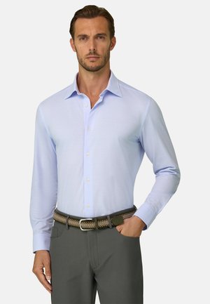 Mann trägt ein hellblaues Hemd mit langen Ärmeln und Knöpfen, graue Hose und einen geflochtenen braunen Gürtel, steht mit einer Hand in der Tasche.