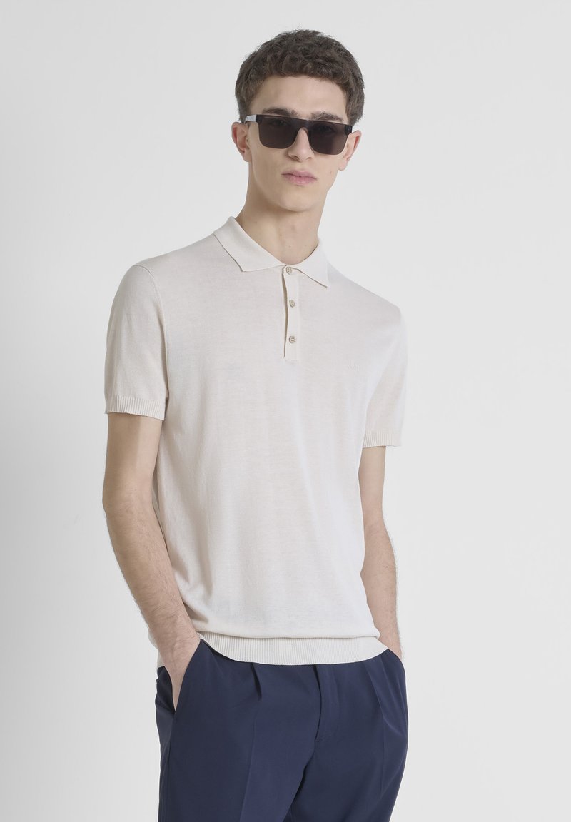 Korte mouwen polo in lichtbeige gebreide stof met kraag en drie knopen. Gecombineerd met donkerblauwe broek, model draagt zwarte zonnebril.