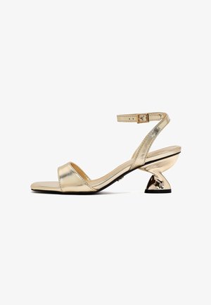Gold metallic heeled sandals feature a slim ankle strap, a wide toe band, and a unique sculptural heel design. Smooth texture and minimal detailing.