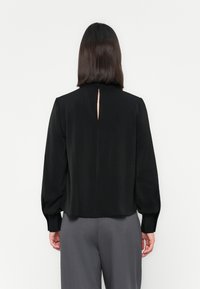 Una donna con i capelli scuri e lisci indossa una blusa nera a maniche lunghe con uno spacco sul retro e pantaloni grigi a vita alta, in piedi contro uno sfondo bianco.