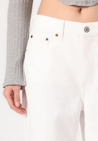 Pantalon en denim blanc à cinq poches, avec des rivets en cuivre et un bouton de fermeture en métal argenté. Texture douce avec une légère élasticité.