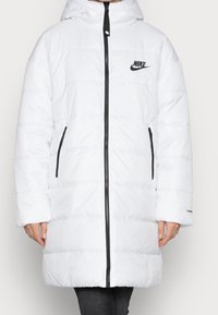Veste longue blanche en duvet avec capuche, fermeture éclair noire, deux poches zippées, logo Nike sur la poitrine, portée sur un pantalon noir.