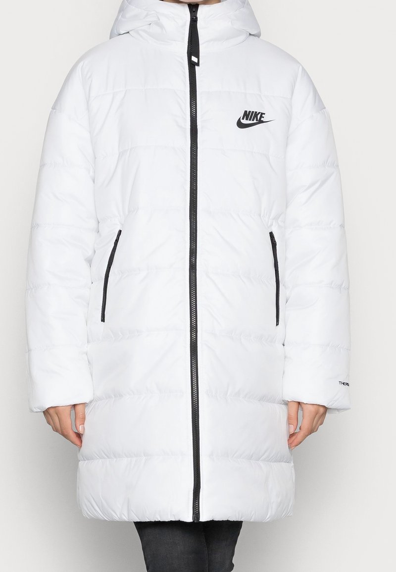 Veste longue blanche en duvet avec capuche, fermeture éclair noire, deux poches zippées, logo Nike sur la poitrine, portée sur un pantalon noir.
