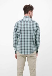 Koszula w kratę z długim rękawem w kolorach zielonym, granatowym i białym; posiada kołnierzyk na guziki, dopasowany krój i zaokrąglony dół. Połączona z beżowymi spodniami.