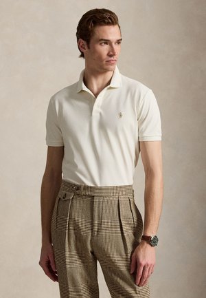 Jonge man die een crème kleurig poloshirt en beige geruite broek met hoge taille draagt, staand tegen een neutrale achtergrond.