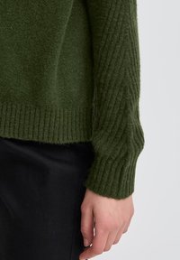 Maglione a maglia verde khaki con texture a coste, caratterizzato da un dettaglio sull'orlo e sui polsini. Materiale morbido e vestibilità rilassata, in contrasto con pantaloni neri.