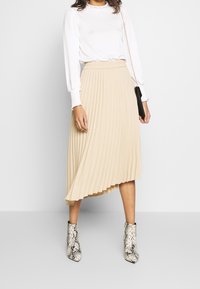 Falda plisada beige con cintura alta elástica, combinada con una blusa blanca de manga larga y botas de tobillo con estampado de serpiente texturizado.