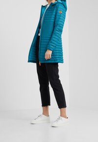 Mujer vistiendo una chaqueta con capucha acolchada en color teal, jeans negros cortos con dobladillos deshilachados y zapatillas blancas, de pie sobre un fondo blanco.