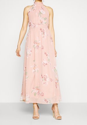 Femme portant une robe maxi rose à fleurs sans manches avec un col montant et des sandales à talons beiges, debout sur un fond blanc.