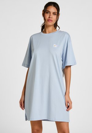 Vrouw die een lichtblauwe oversized T-shirtjurk draagt met een klein wit en geel madeliefje geborduurd op de borst, staand tegen een effen achtergrond.