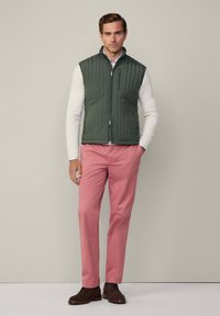 Grüne gesteppte Weste über einem weißen Pullover, kombiniert mit rosafarbenen Hosen und braunen Schuhen. Die Weste hat einen Stehkragen und einen Reißverschluss.