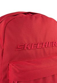 Mochila roja con una tela texturizada. Cuenta con un cierre superior de cremallera y un bolsillo frontal. "SKECHERS" está embellecido en rojo a juego en la parte frontal.