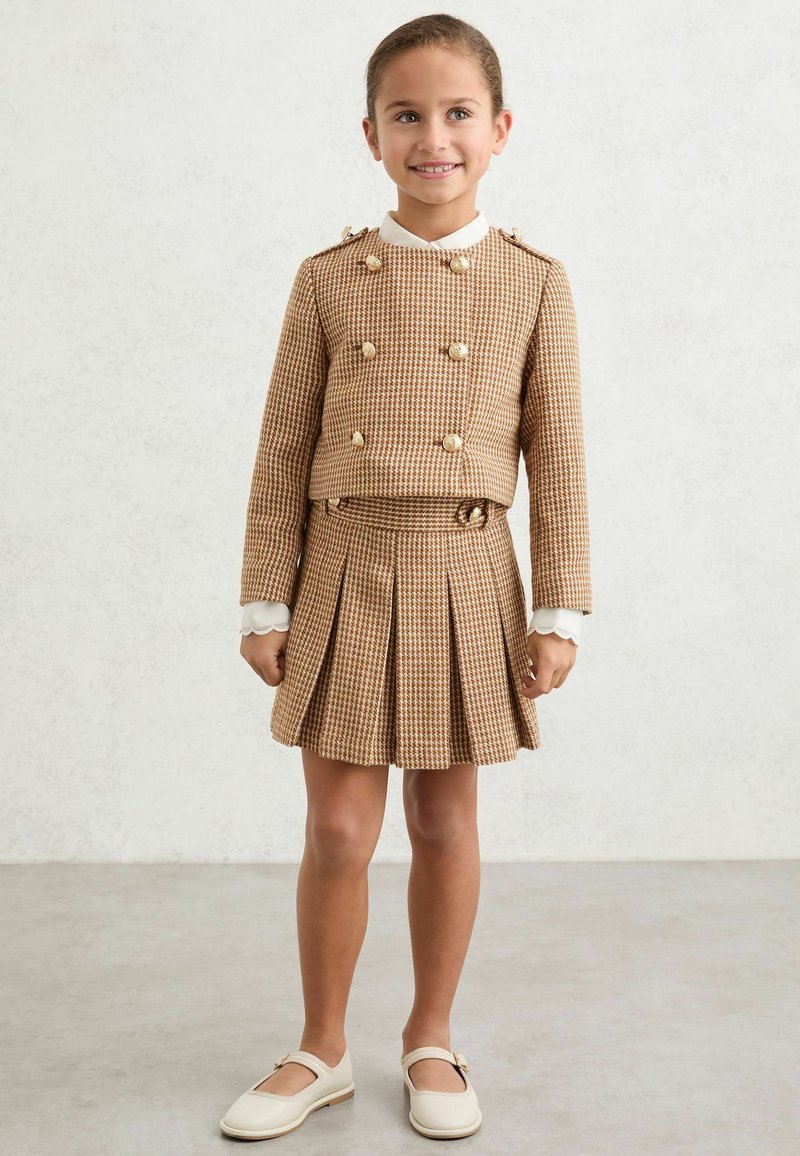 Noor tüdruk naeratab, kandes pruuni houndstooth-mustriga kahekordse nööpidega jopet ja sobiva voldilise seelikut valge krae ja kingadega.