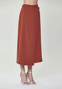 Jupe midi de couleur rouille en tissu doux, présentant un design asymétrique et un accent décoratif en argent au niveau de la taille.