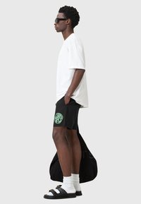 Pantalones cortos negros con un logotipo circular verde, combinados con una camiseta blanca de gran tamaño. Los accesorios incluyen gafas de sol negras y sandalias negras con calcetines.