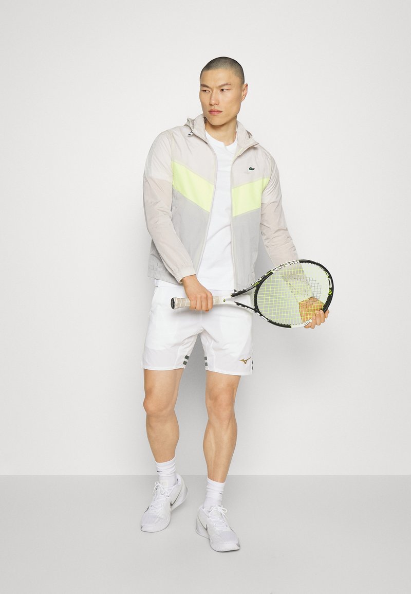 Lichtgewicht, grijs en geel sportjack met capuchon, gecombineerd met een wit shirt en witte shorts. Houdt een tennisracket vast. Witte schoenen.