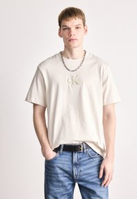 Μπεζ βαμβακερό μπλουζάκι με στρογγυλή λαιμόκοψη και κοντά μανίκια, που διαθέτει το ανάγλυφο λογότυπο "cK" στο κέντρο. Συνδυάζεται με μπλε τζιν παντελόνι και μαύρη ζώνη.