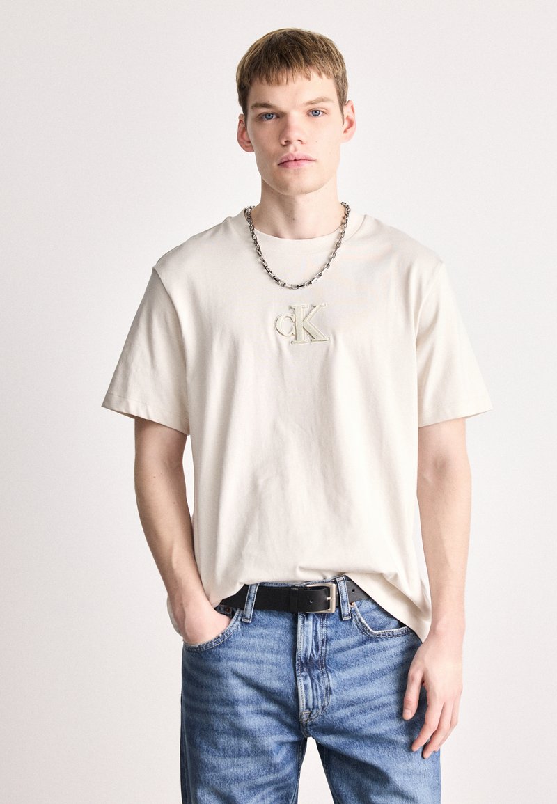 Μπεζ βαμβακερό μπλουζάκι με στρογγυλή λαιμόκοψη και κοντά μανίκια, που διαθέτει το ανάγλυφο λογότυπο "cK" στο κέντρο. Συνδυάζεται με μπλε τζιν παντελόνι και μαύρη ζώνη.