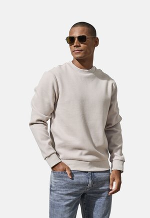 Mann trägt strukturiertes beiges Sweatshirt, hellblaue Jeans und Sonnenbrille, steht mit einer Hand in der Tasche vor einfarbigem Hintergrund.