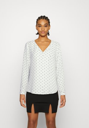 White long-sleeve blouse with navy polka dots, featuring a V-neckline and a slightly curved hem. Paired with a black skirt with slits.
