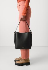 Sac fourre-tout en cuir noir avec une texture, matériel doré et logo. Forme arrondie, tenu par une main, associé à un pantalon rose et des chaussures marron.