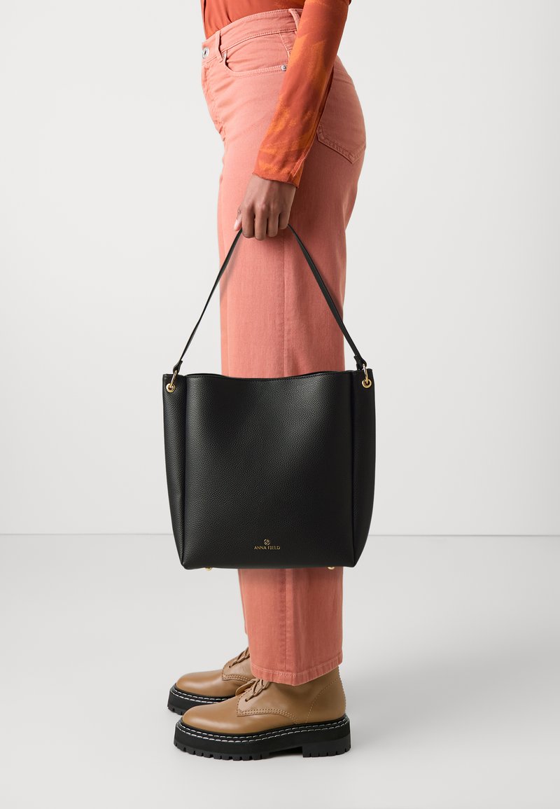 Sac fourre-tout en cuir noir avec une texture, matériel doré et logo. Forme arrondie, tenu par une main, associé à un pantalon rose et des chaussures marron.