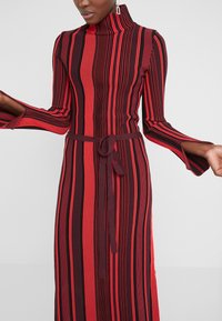 Femme portant une robe à manches longues et col haut avec des rayures verticales rouges et noires et une ceinture nouée à la taille.