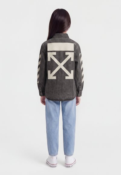 Άτομο με μακριά σκούρα μαλλιά που φοράει μαύρο τζιν μπουφάν με κρεμ βέλη στην πλάτη και τα μανίκια, ανοιχτό μπλε τζιν και λευκά αθλητικά παπούτσια.