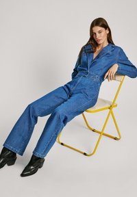 Denim kombinezon v srednji modri barvi z dolgimi rokavi, ovratnikom in pasom. Nosi črne čevlje na višjem petniku, sedeč na rumeni stolu.