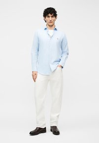 Άνδρας που φορά πουκάμισο με ανοιχτόμπλε ρίγες και κουμπιά, λευκό παντελόνι και μαύρα παπούτσια, στέκεται με το ένα χέρι στην τσέπη μπροστά σε λευκό φόντο.