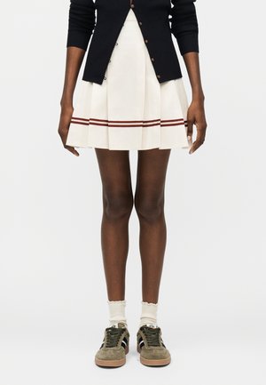 Personne portant une jupe plissée crème avec des rayures marron, un cardigan noir, des chaussettes blanches et des baskets en daim vert, debout devant un fond blanc.