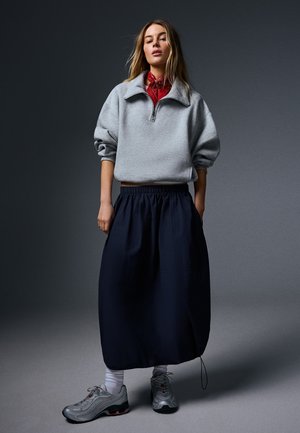 Femme portant un sweat-shirt gris à fermeture éclair quart, une écharpe rouge, une longue jupe foncée, des chaussettes blanches et des baskets grises sur un fond gris foncé.