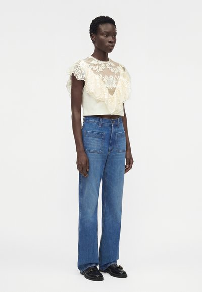 Γυναίκα που στέκεται, φοράει ένα κρεμ δαντελωτό τοπ με βολάν, ψηλόμεσο μπλε τζιν με μπροστινές τσέπες και μαύρα μοκασίνια σε απλό λευκό φόντο.