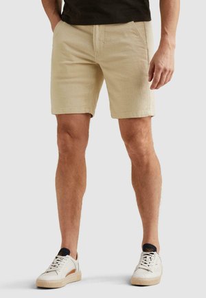 Mann trägt beige strukturierte Shorts, schwarzes T-Shirt und weiße Turnschuhe, steht vor einem schlichten hellen Hintergrund.