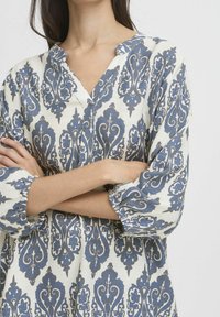 Weiße Tunika mit blauem Paisleymuster, mit V-Ausschnitt, dreiviertel Ärmeln und gerafften Manschetten. Weiche Textur, leichtes Material.