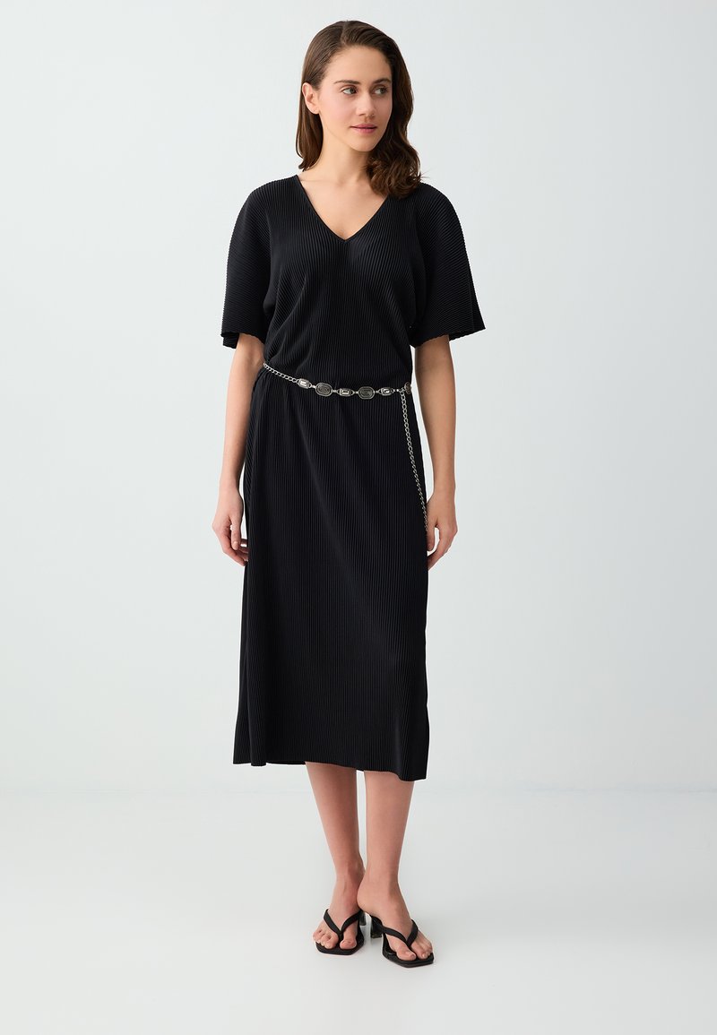 Femme debout portant une robe midi noire côtelée à manches courtes, une ceinture chaîne argentée et des sandales noires à talons.
