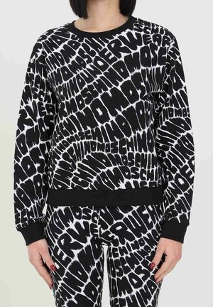 Kobieta ubrana w dopasowaną czarno-białą bluzę i spodnie z wyrazistym, zdeformowanym wzorem tekstowym powtarzającym słowa "MOSCHINO" i "WATERS".