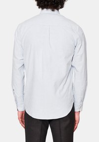 Mężczyzna w jasnoniebieskiej, białej koszuli w cienkie paski z długim rękawem oraz ciemnych spodniach, pokazany od tyłu na jednolitym tle.