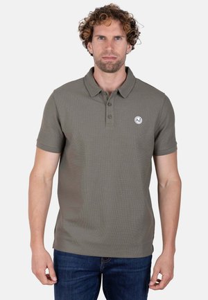Hombre con pelo rizado que lleva un polo de manga corta verde oliva y vaqueros azules, de pie contra un fondo liso y claro.